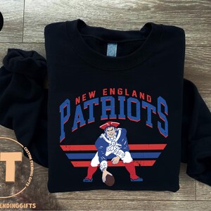 May include: A black long-sleeve sweatshirt featuring a graphic of a football player in a red, white, and blue uniform. The text "NEW ENGLAND PATRIOTS" is printed in blue above the player, with red and blue horizontal stripes below the figure.