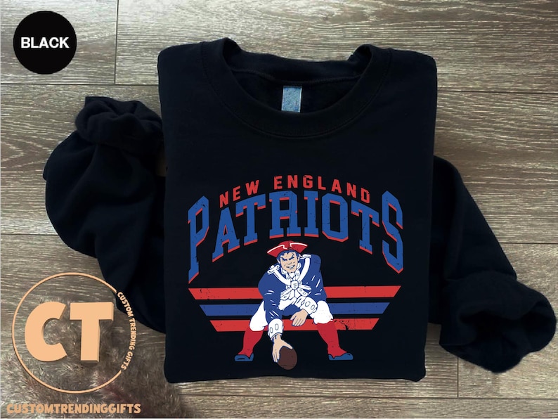 May include: A black long-sleeve sweatshirt featuring a graphic of a football player in a red, white, and blue uniform. The text "NEW ENGLAND PATRIOTS" is printed in blue above the player, with red and blue horizontal stripes below the figure.