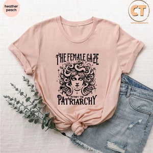 K&ouml;nnte beinhalten: Ein rosa T-Shirt mit einer schwarzen Grafik einer Frau mit Schlangen als Haaren. Der Text auf dem Shirt lautet "The Female Gaze Petrified the Patriarchy."