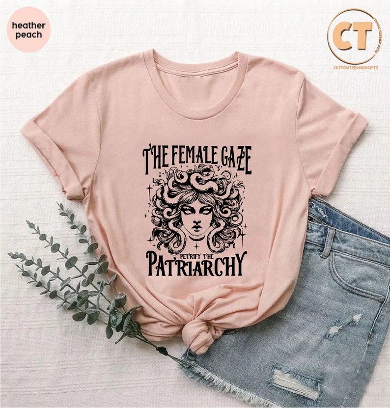 K&ouml;nnte beinhalten: Ein rosa T-Shirt mit einer schwarzen Grafik einer Frau mit Schlangen als Haaren. Der Text auf dem Shirt lautet "The Female Gaze Petrified the Patriarchy."