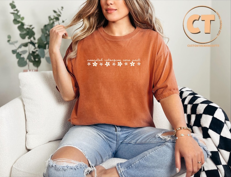 K&ouml;nnte beinhalten: Eine Frau tr&auml;gt ein rostfarbenes T-Shirt mit dem Text "neonatal intensive care unit" und wei&szlig;en Blumen, die auf die Vorderseite gedruckt sind. Sie sitzt auf einem wei&szlig;en Sofa mit einer schwarz-wei&szlig;en karierten Wolldecke.