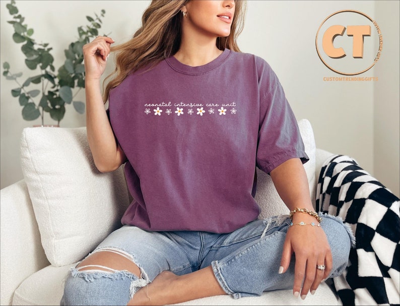 K&ouml;nnte beinhalten: Ein lila T-Shirt mit dem Text "neonatal intensive care unit" und einer Reihe kleiner wei&szlig;er Blumen.