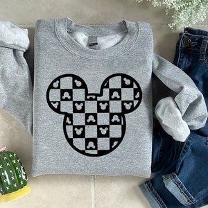 May include: A gray sweatshirt featuring a black and white checkered Mickey Mouse head design. The design incorporates Mickey Mouse silhouettes within the checkered pattern. The sweatshirt is displayed with blue jeans and a decorative cactus.