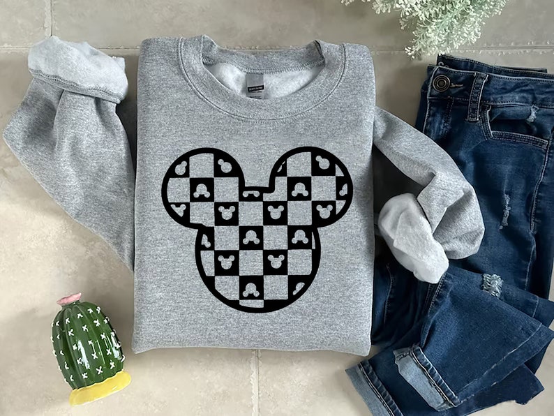 May include: A gray sweatshirt featuring a black and white checkered Mickey Mouse head design. The design incorporates Mickey Mouse silhouettes within the checkered pattern. The sweatshirt is displayed with blue jeans and a decorative cactus.