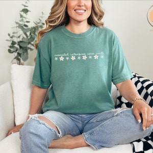 K&ouml;nnte beinhalten: Ein blaugr&uuml;nes T-Shirt mit dem Text "neonatal intensive care unit" und einer Reihe wei&szlig;er G&auml;nsebl&uuml;mchen mit gelben Zentren.
