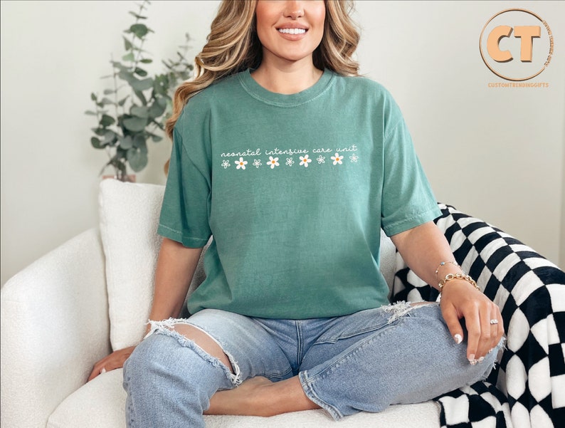 K&ouml;nnte beinhalten: Ein blaugr&uuml;nes T-Shirt mit dem Text "neonatal intensive care unit" und einer Reihe wei&szlig;er G&auml;nsebl&uuml;mchen mit gelben Zentren.