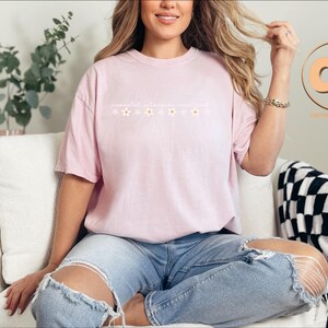 K&ouml;nnte beinhalten: Ein pinkfarbenes T-Shirt mit einem Blumendesign und dem Text "remarkable ordinary you". Das Model tr&auml;gt das Shirt mit einer blauen Jeans.