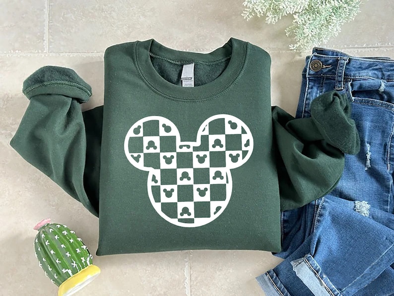 May include: Dark green sweatshirt featuring a white Mickey Mouse head design with a checkered pattern. The design incorporates Mickey Mouse silhouettes within the checks. The sweatshirt is folded, with the sleeves rolled up, and is paired with blue jeans.