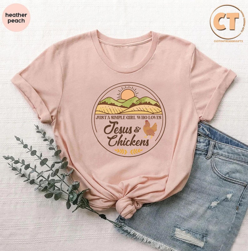 K&ouml;nnte beinhalten: Ein rosa T-Shirt mit einem braunen Kreis-Motiv, das einen Sonnenuntergang &uuml;ber einem gr&uuml;nen Feld, ein Huhn und den Text "Just a simple girl who loves Jesus & Chickens" zeigt.