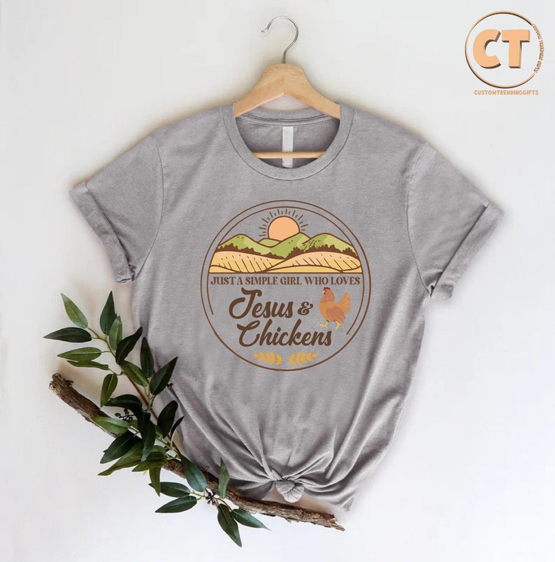K&ouml;nnte beinhalten: Ein graues T-Shirt mit einem braunen Kreis-Motiv. Das Motiv zeigt einen Sonnenuntergang &uuml;ber einem Feld, ein Huhn und den Text "Just a simple girl who loves Jesus & Chickens".