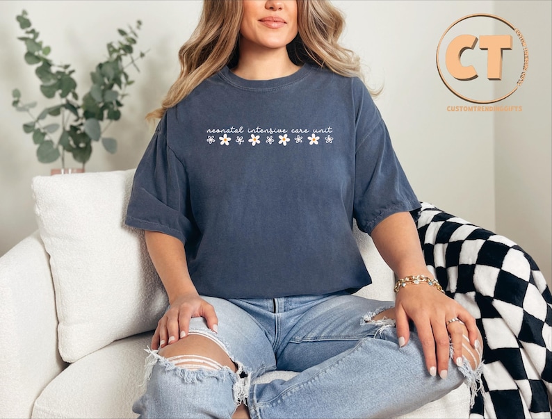 K&ouml;nnte beinhalten: Ein marineblaues T-Shirt mit dem Text "neonatal intensive care unit" und einer Reihe wei&szlig;er Blumen, die auf der Vorderseite aufgedruckt sind. Das Shirt wird von einer Person getragen, die auf einem wei&szlig;en Sofa sitzt.