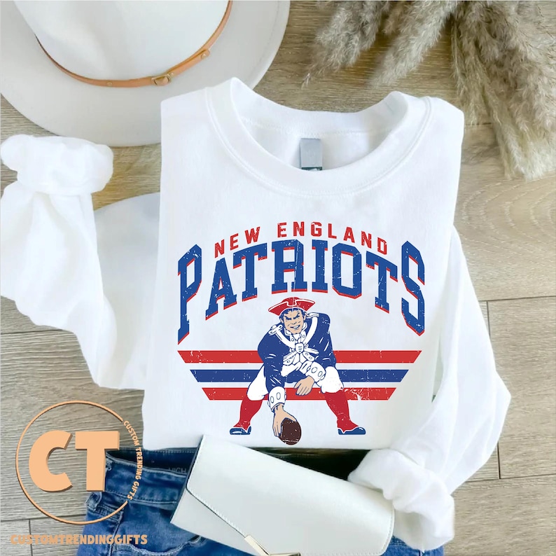 May include: White crew neck sweatshirt featuring the words "NEW ENGLAND PATRIOTS" in blue and red block letters. Below the text is a graphic of a colonial-era man in a blue coat and red pants holding a football. The sweatshirt is folded.