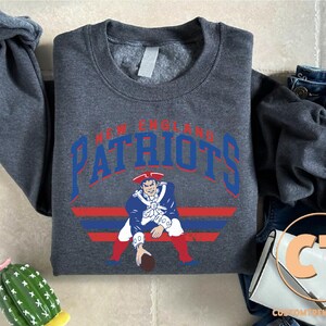 May include: A dark charcoal gray crewneck sweatshirt featuring the text New England Patriots in blue and red lettering above a graphic of a football player in a blue, white, and red uniform, positioned above horizontal blue and red stripes.