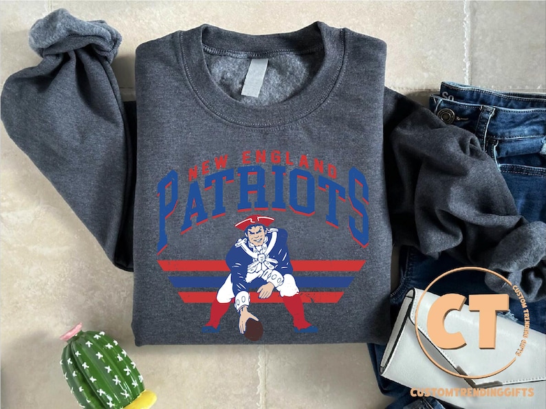 May include: A dark charcoal gray crewneck sweatshirt featuring the text New England Patriots in blue and red lettering above a graphic of a football player in a blue, white, and red uniform, positioned above horizontal blue and red stripes.