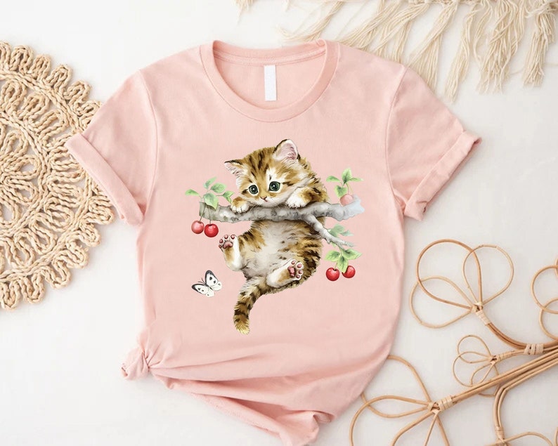 May include: A pink t-shirt with a watercolor illustration of a brown and white kitten hanging from a branch with red cherries. A white butterfly is flying near the kitten.