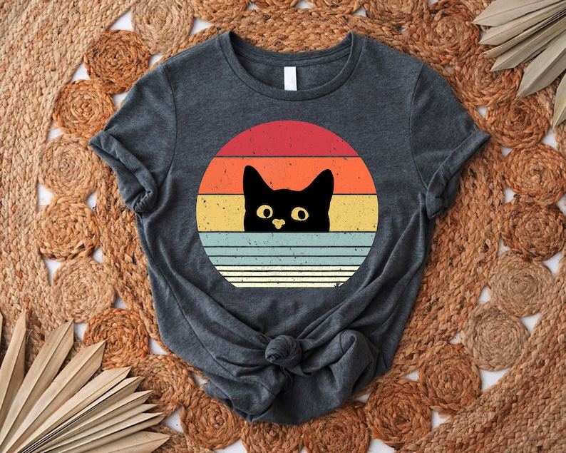 May include: A dark gray t-shirt with a retro-style sunset graphic featuring a black cat peeking out from behind the stripes.