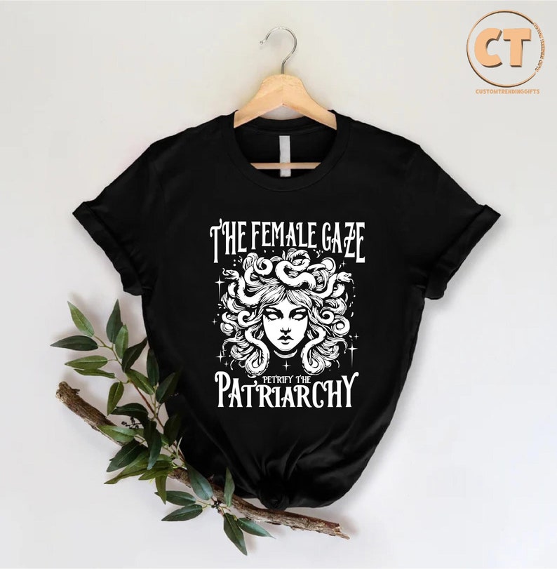 K&ouml;nnte beinhalten: Schwarzes T-Shirt mit einer wei&szlig;en Grafik einer Frau mit Schlangen als Haaren. Der Text "The Female Gaze" und "Petrify the Patriarchy" ist auf dem Shirt aufgedruckt.