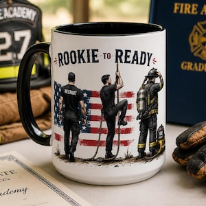 Puede incluir: Una taza de cerámica blanca con asa e interior negros, que presenta el texto ROOKIE TO READY y una ilustración de tres figuras frente a una bandera estadounidense, mostrando a un recluta, una persona trepando una cuerda y un bombero.