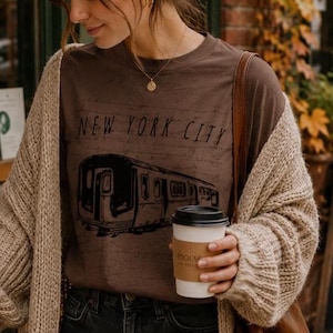 May include: A brown short-sleeved t-shirt featuring a black graphic of a subway train and the text NEW YORK CITY, worn under a chunky beige knit cardigan. The shirt is paired with black denim jeans and a brown leather shoulder bag.