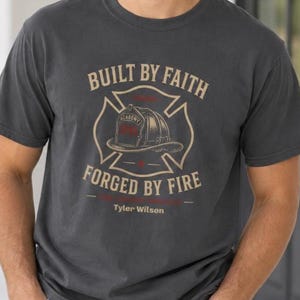 Puede incluir: Una camiseta gris carbón de manga corta con un gráfico de un casco de bombero dentro de un emblema de cruz de Malta. El texto dice BUILT BY FAITH FORGED BY FIRE e incluye el nombre Tyler Wilson impreso en tonos beige claro y rojo.