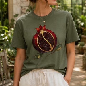 Puede incluir: Una camiseta de manga corta color verde oliva con un estampado gráfico de una granada con estética de vidriera. La granada está representada en tonos rojo oscuro y morado con semillas visibles y hojas verdes.