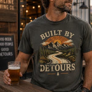 May include: A charcoal gray short-sleeve t-shirt featuring a graphic print of a winding road leading toward snow-capped mountains under a rising sun. The text BUILT BY DETOURS is printed in a vintage-style font above and below the mountain scene.