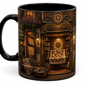 May include: A black ceramic mug featuring a detailed illustration of a storefront labeled Mandie's Quilt Shop. The scene depicts a cozy shop interior with hanging quilts, a sleeping cat in a basket, and exterior signage reading Quilt Shop and Stitching Memories One Quilt at a Time.