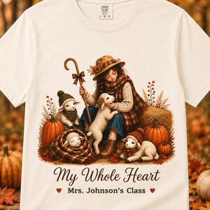 May include: A cream-colored short-sleeve t-shirt featuring a graphic of a person in a hat and scarf with three lambs, pumpkins, and autumn foliage. The text reads "My Whole Heart" and "Mrs. Johnson’s Class" with two small red heart icons.