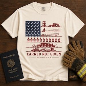 Puede incluir: Una camiseta de manga corta color crema con un gráfico de la bandera estadounidense compuesto por imágenes de bomberos, incluyendo una estación, un bombero con manguera, un camión escalera y una fila de bomberos, con el texto EARNED NOT GIVEN.