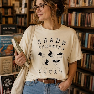 May include: A cream-colored short-sleeve t-shirt featuring black text that reads SHADE THROWING SQUAD and various black hand shadow puppet illustrations including a rabbit, a bird, and a dog, paired with blue denim jeans and a brown leather belt.