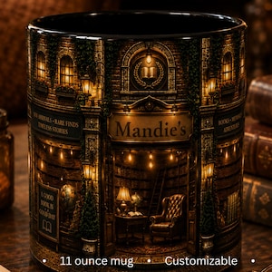 May include: A black ceramic mug featuring a detailed wrap-around illustration of a bookstore interior with warm lighting, a leather armchair, and bookshelves. The text Mandie's is printed above the entrance, with A GOOD BOOK IS A GOOD DAY below.