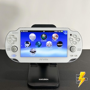 May include: A white Sony PS Vita handheld gaming console is displayed on a black stand. The screen shows various game icons, including DS Vita, Hollow Knight, and Assassin's Creed. The console is in a black stand with the text "KAKUSIGA" on the base.