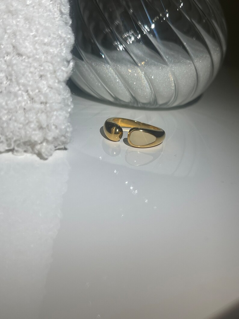 Peut inclure: Une bague en or avec une pierre ovale blanche. La bague est ouverte d'un c&ocirc;t&eacute; et a une boule ronde en or de l'autre c&ocirc;t&eacute;.