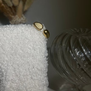 Peut inclure: Une paire de boucles d'oreilles en or avec une pierre ovale blanche. Les boucles d'oreilles sont sur une surface blanche et duveteuse.