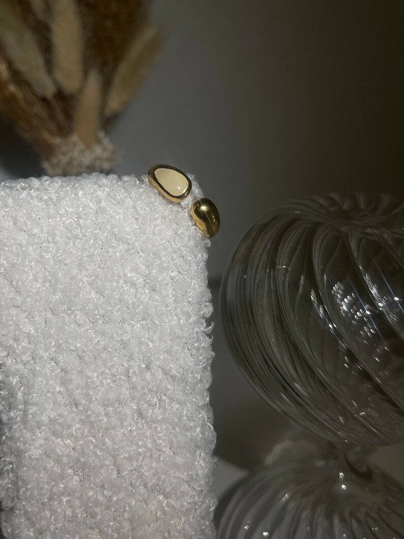 Peut inclure: Une paire de boucles d'oreilles en or avec une pierre ovale blanche. Les boucles d'oreilles sont sur une surface blanche et duveteuse.