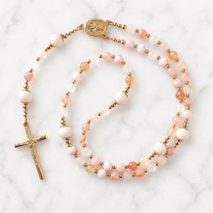 May include: A rosary featuring translucent pink and peach-toned gemstone beads interspersed with smooth white beads and small gold-toned metal spacers. The piece includes a central gold-toned medal and a gold-toned crucifix pendant at the end.