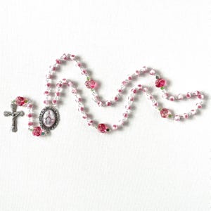 May include: A rosary featuring pink and white rose-shaped beads interspersed with small green accent beads. The piece includes a silver-toned metal crucifix and an oval-shaped centerpiece medallion depicting a figure in a white and blue robe.