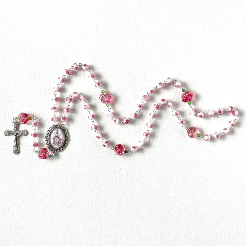 May include: A rosary featuring pink and white rose-shaped beads interspersed with small green accent beads. The piece includes a silver-toned metal crucifix and an oval-shaped centerpiece medallion depicting a figure in a white and blue robe.