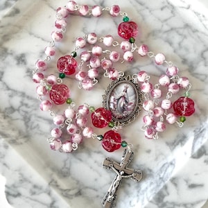 May include: A rosary featuring white beads with pink rose patterns, translucent red rose-shaped beads, and small green accent beads. The rosary includes a silver-toned metal crucifix and an oval centerpiece depicting a figure in a flowing robe.