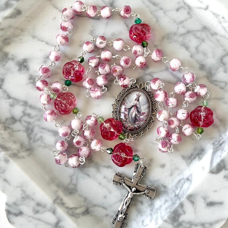 May include: A rosary featuring white beads with pink rose patterns, translucent red rose-shaped beads, and small green accent beads. The rosary includes a silver-toned metal crucifix and an oval centerpiece depicting a figure in a flowing robe.