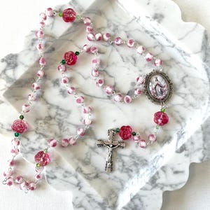 May include: A rosary featuring pink and white marbled beads interspersed with translucent dark pink rose-shaped beads and small green crystals. The rosary includes a silver-toned metal crucifix and an oval centerpiece depicting a religious figure.