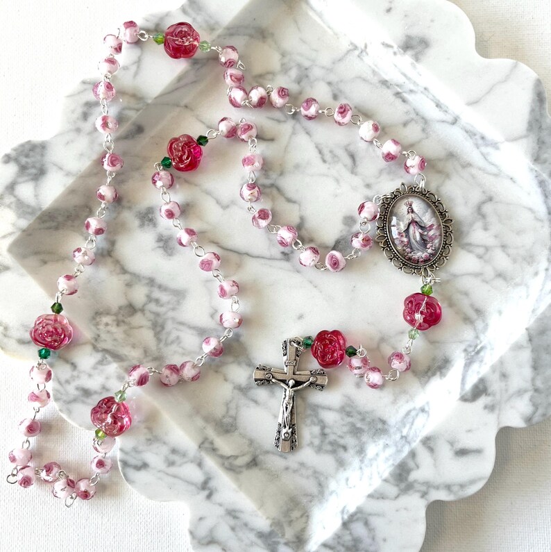 May include: A rosary featuring pink and white marbled beads interspersed with translucent dark pink rose-shaped beads and small green crystals. The rosary includes a silver-toned metal crucifix and an oval centerpiece depicting a religious figure.