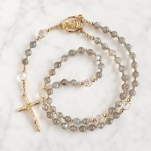 May include: A rosary featuring grey labradorite beads and clear faceted crystal beads, accented with gold-toned metal spacers. The piece includes a gold-toned crucifix and a central medal depicting a profile of a figure, all linked by gold wire.
