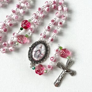 May include: A rosary featuring light pink beads with white rose patterns, interspersed with translucent dark pink rose-shaped beads and small green crystals. The centerpiece is an oval metal pendant depicting a figure, connected to a silver-tone crucifix.