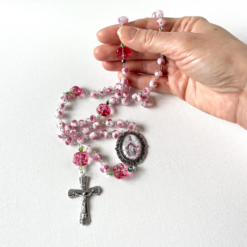 May include: A rosary featuring pink and white floral patterned beads, translucent pink rose-shaped beads, and small green accent beads. The rosary includes a silver-toned metal crucifix and an oval-shaped centerpiece depicting a religious figure.