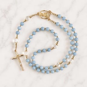 May include: A rosary with light blue beads and gold-colored accents. The rosary features a gold crucifix and a gold centerpiece with a religious figure. Clear crystal beads are interspersed throughout the design.