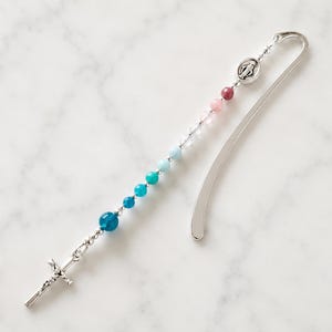 May include: A beaded bookmark with a silver cross and a Virgin Mary medallion. The beads transition in color from blue to pink. The bookmark is made of metal and has a curved design.