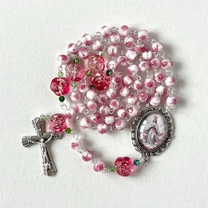 May include: A rosary featuring pink and white rose-shaped beads, small green accent beads, and a silver-toned metal crucifix. An oval-shaped pendant depicts a figure in a white robe with a crown, framed by an ornate silver-toned metal border.