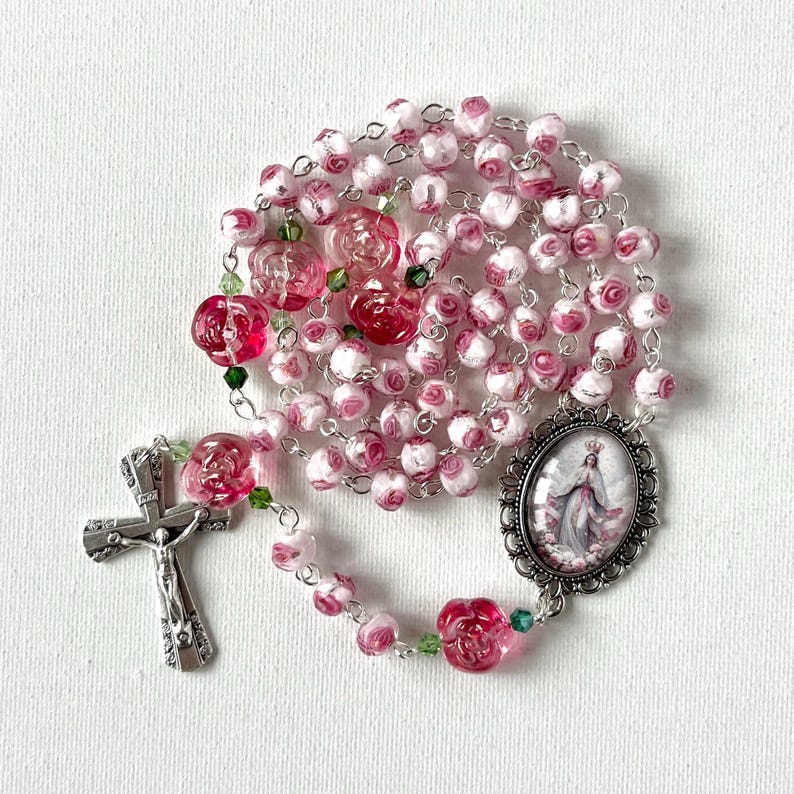 May include: A rosary featuring pink and white rose-shaped beads, small green accent beads, and a silver-toned metal crucifix. An oval-shaped pendant depicts a figure in a white robe with a crown, framed by an ornate silver-toned metal border.