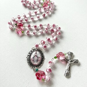 May include: A rosary featuring pink and white rose-shaped beads connected by silver-toned metal links. The piece includes a silver-toned crucifix and an oval-shaped centerpiece medallion depicting a figure in a robe, accented with small green beads.
